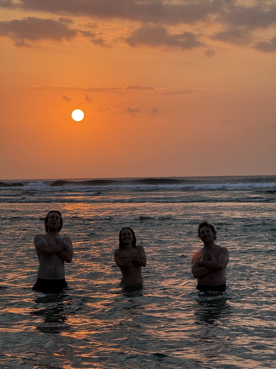 Trois personnes debout dans l'eau, face au soleil qui se couche sur l'océan