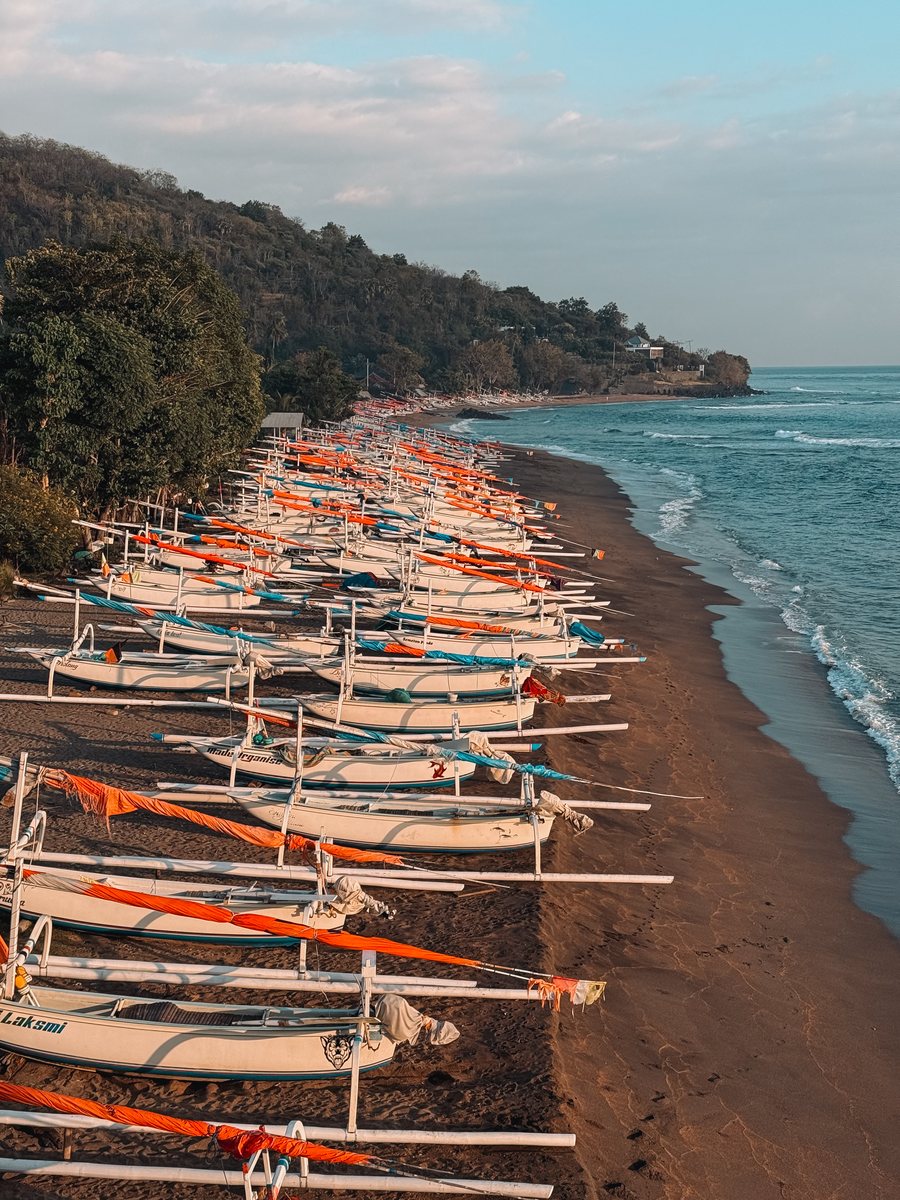 Des dizaines de bateaux de pêche traditionnels alignés sur une plage de sable sombre, au lever du jour