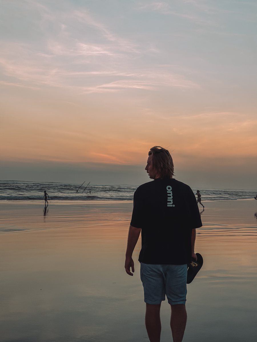 Julien de dos sur la plage de Canggu, une planche à la main, face au coucher de soleil