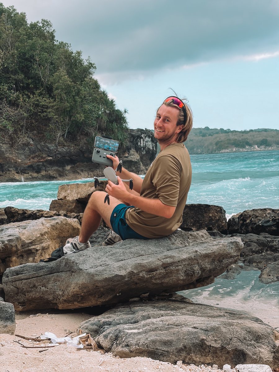 Julien assis sur des rochers au bord de l'eau, en train de piloter un drone