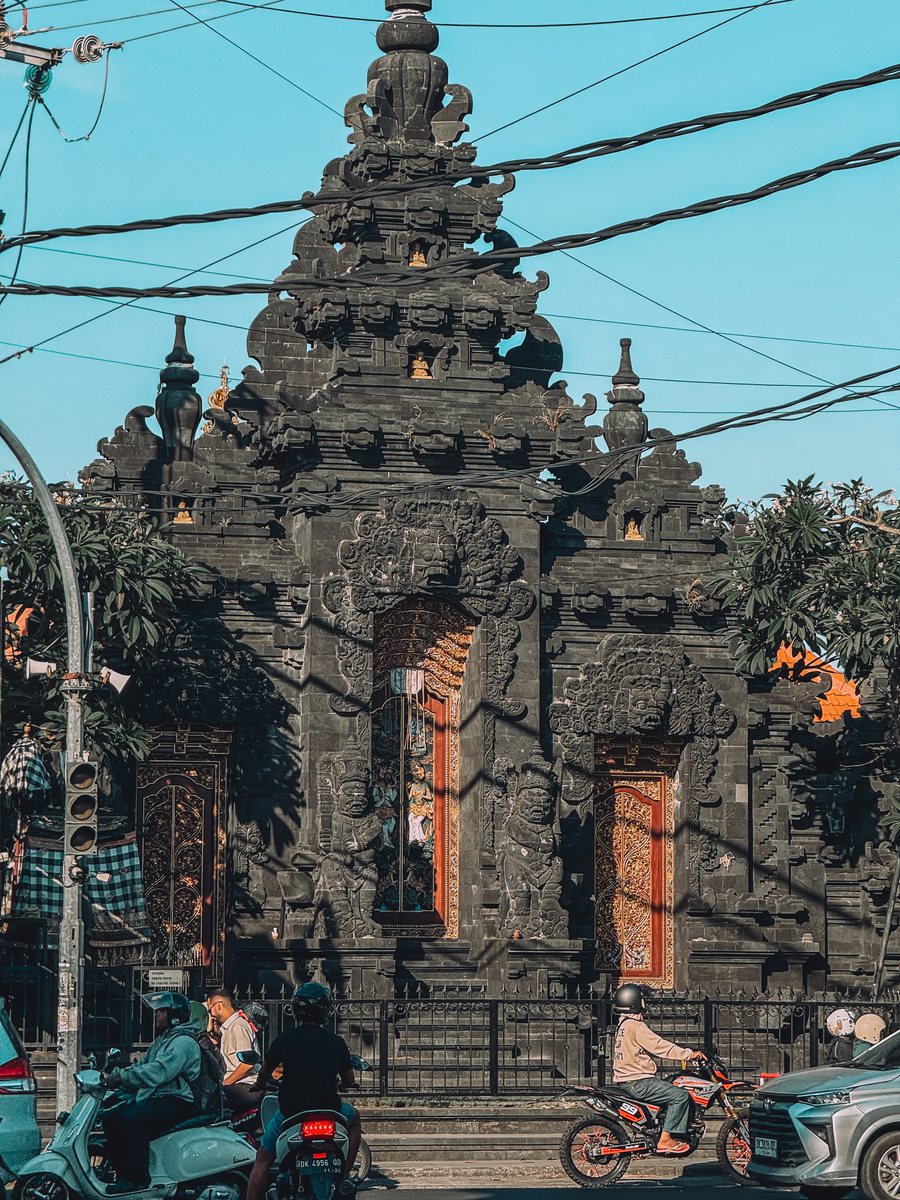 Un portail de temple en pierre sculptée au bord d'une route, entre les câbles électriques et les scooters