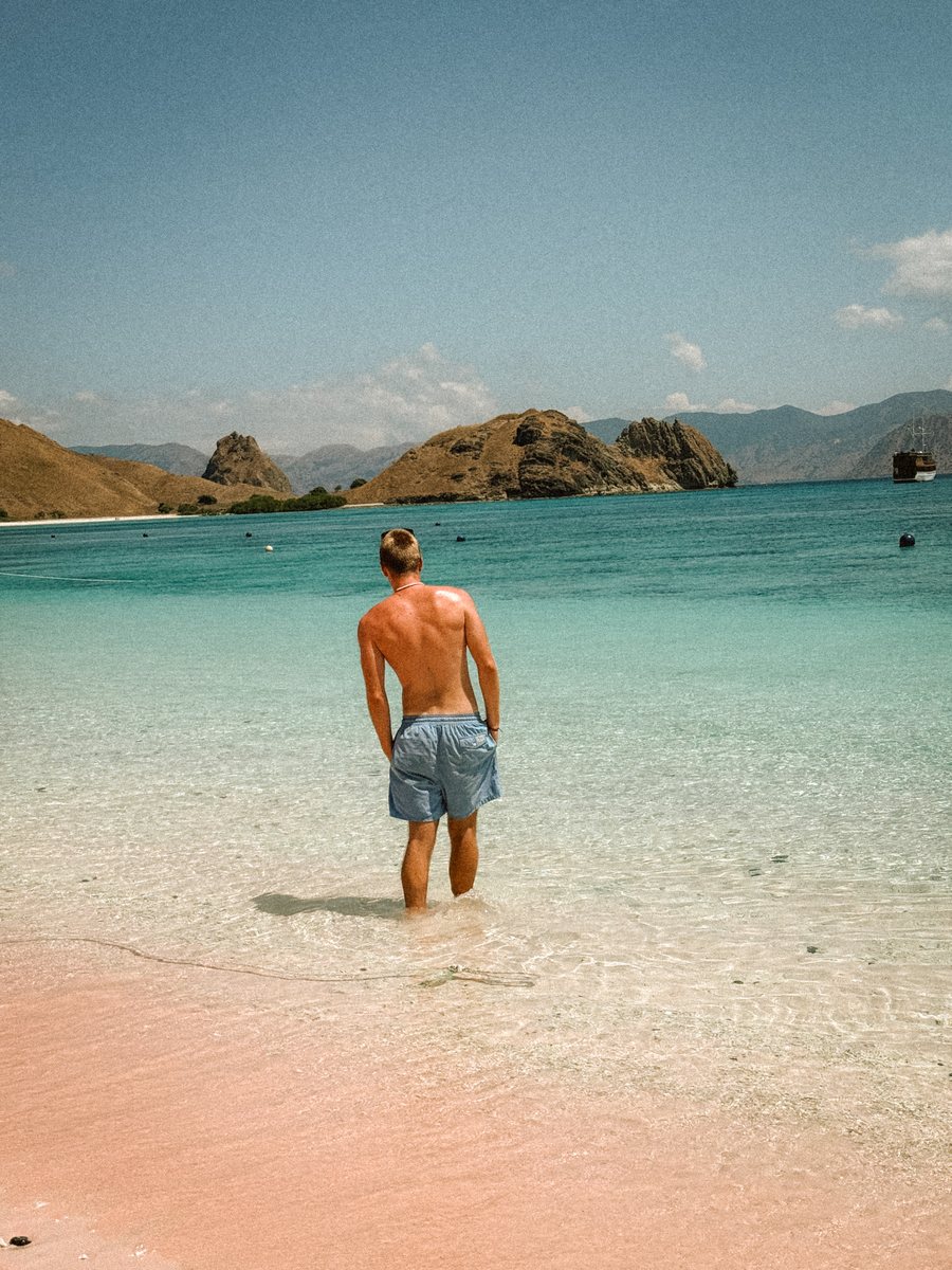 Julien entrant dans une eau turquoise depuis une plage au sable légèrement rosé