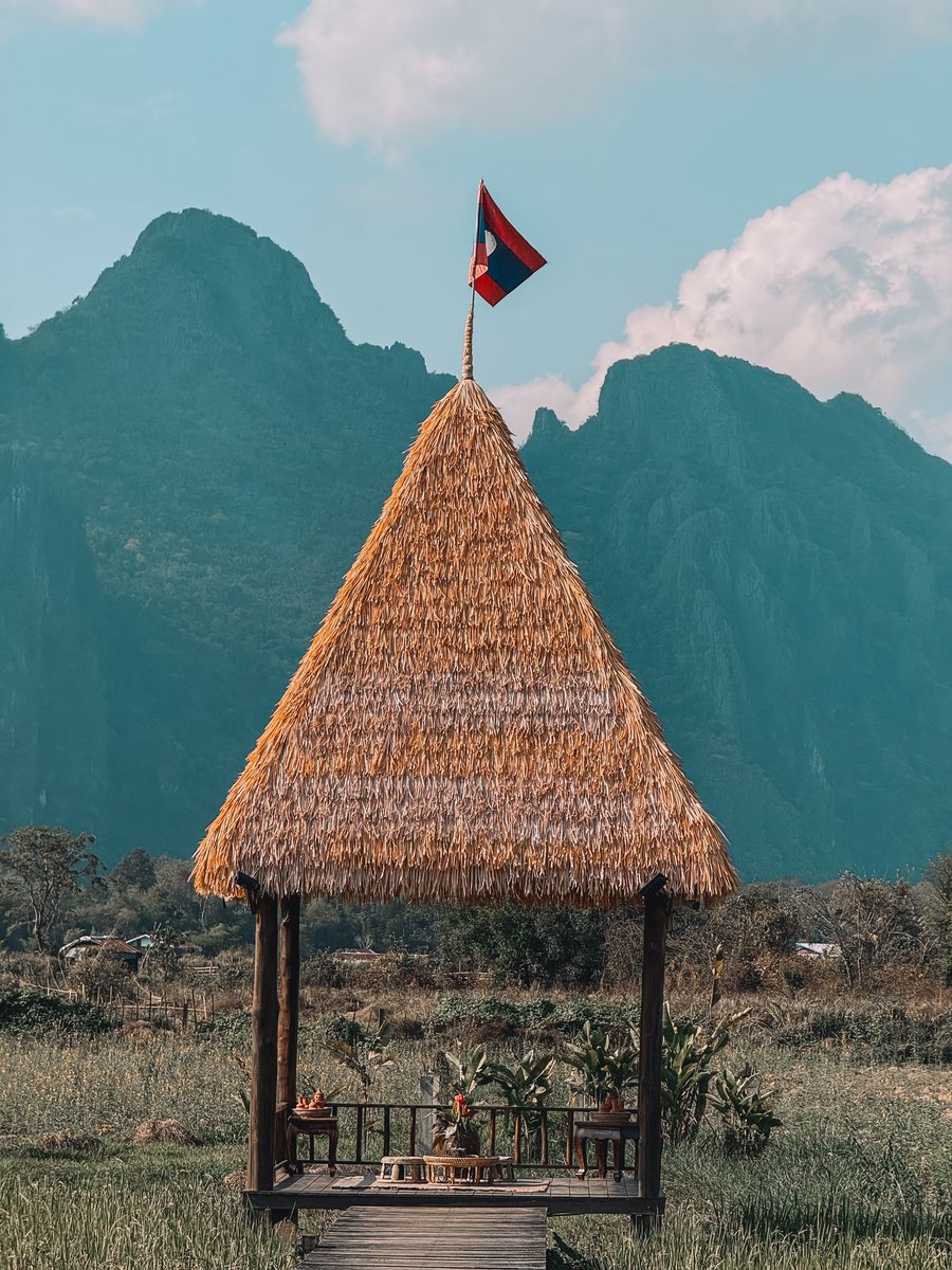Kiosque au toit de paille surmonté du drapeau laotien, devant des montagnes calcaires