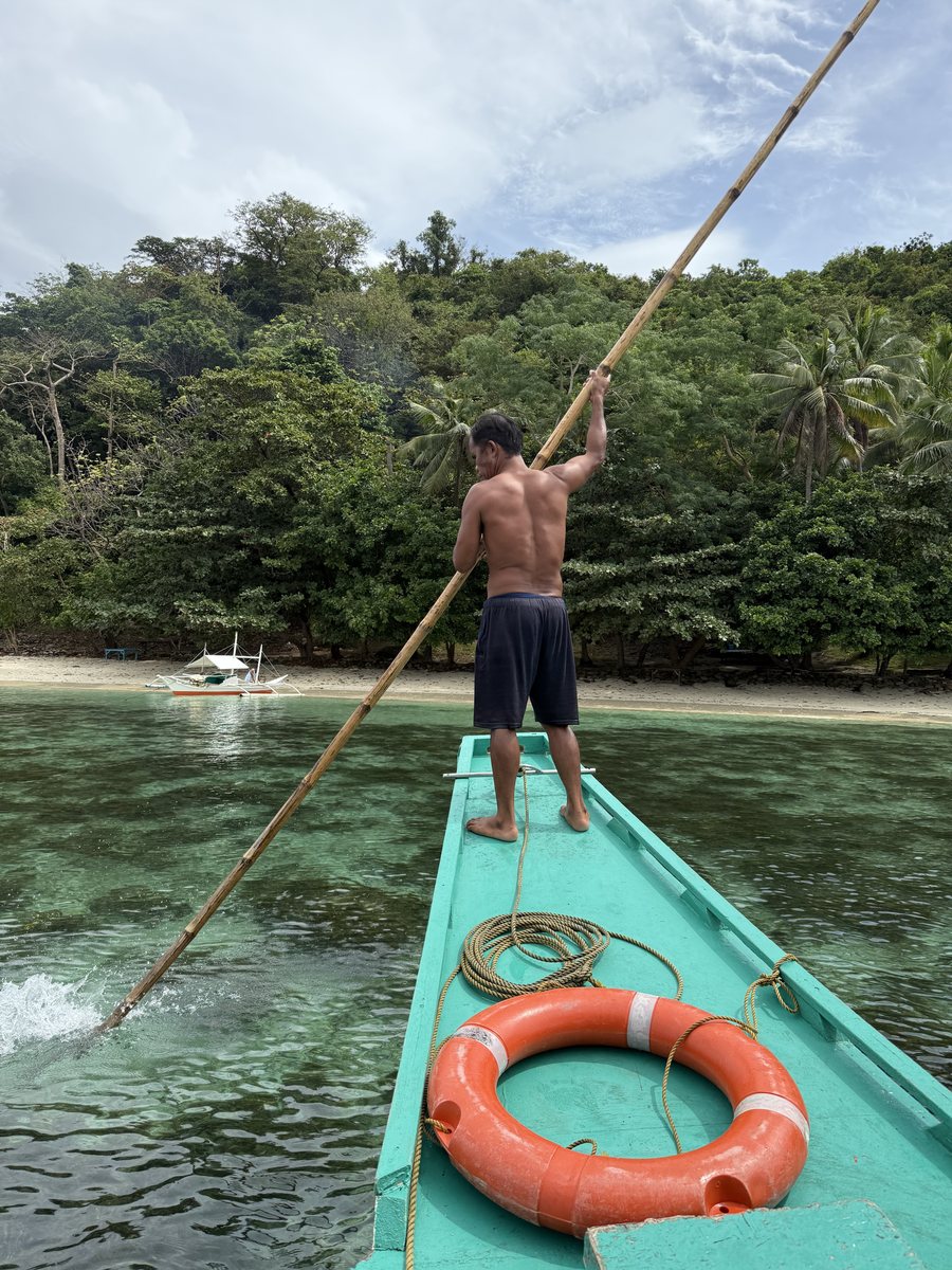 Marin debout à la proue d'un bangka, la perche à la main, devant une île boisée
