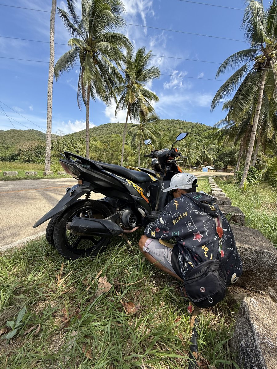 Scooter garé au bord d'une route, sacs à dos posés contre la selle, cocotiers en fond