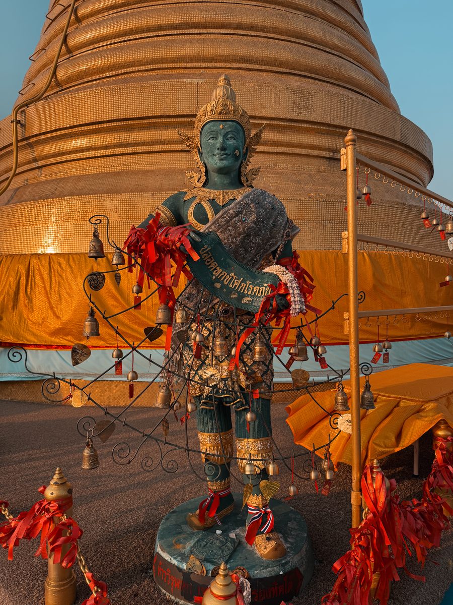 Statue et clochettes couvertes de rubans rouges devant un chedi doré, à Bangkok