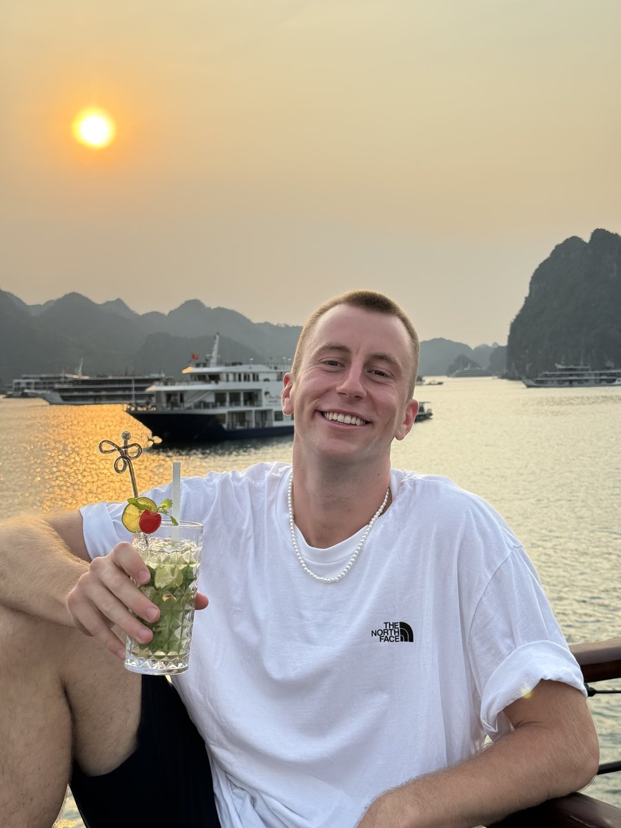Julien un verre à la main sur le pont d'un bateau, au coucher du soleil dans la baie